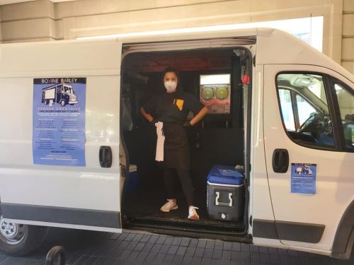 A person in a mask stands in the doorway of a van, ready to serve frozen cocktails, with a menu displayed on the side.