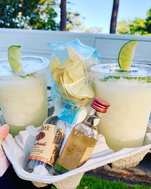 Two cups of cocktails with lime wedges, a bag of chips, and a small bottle of vodka on a tray outdoors.