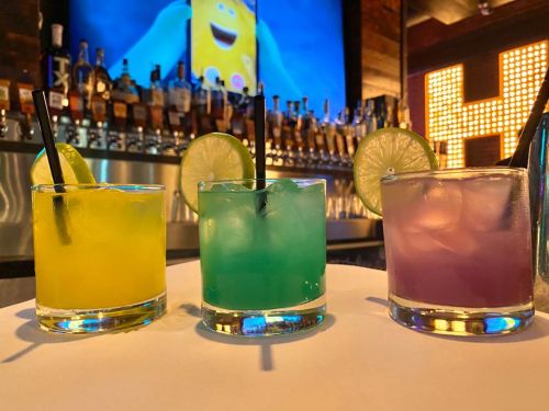 Three colorful cocktails with lime slices on a bar, with a vibrant backdrop of bottles and a screen.