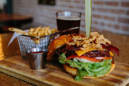 A gourmet burger with bacon, cheese, lettuce, and tomato, served with fries and a pint of beer on a wooden board.