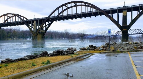 A misty river scene featuring a large, arched bridge and rocky shoreline under a cloudy sky.