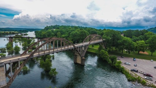 Aerial view of a curved bridge over a river, surrounded by lush greenery and a cloudy sky.
