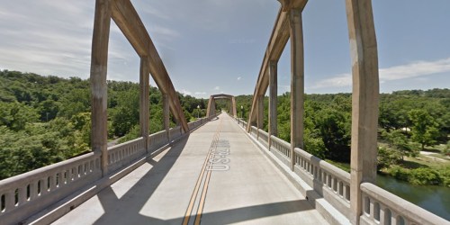 A wide view of a concrete bridge with arched supports, surrounded by lush greenery and a clear blue sky.