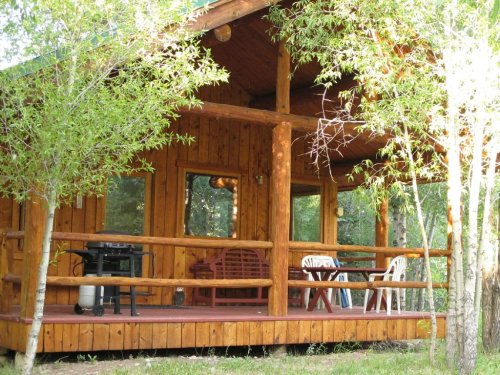 A cozy wooden cabin with a porch, surrounded by trees, featuring chairs and a grill.