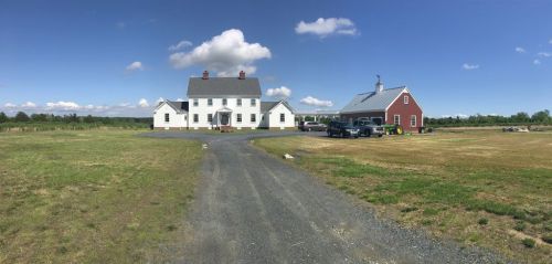 A large white farmhouse with a gray roof, next to a red barn, set in a grassy field under a blue sky with clouds.