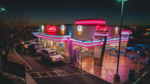 A brightly lit diner with neon signs, cars parked outside, and outdoor seating under a twilight sky.