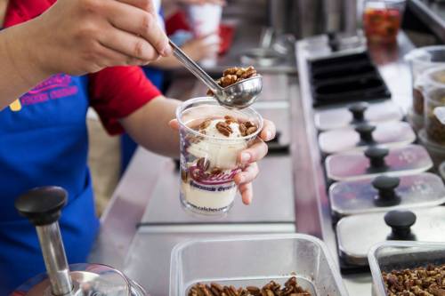 A person scoops nuts onto a cup of ice cream at a dessert counter, surrounded by various toppings.