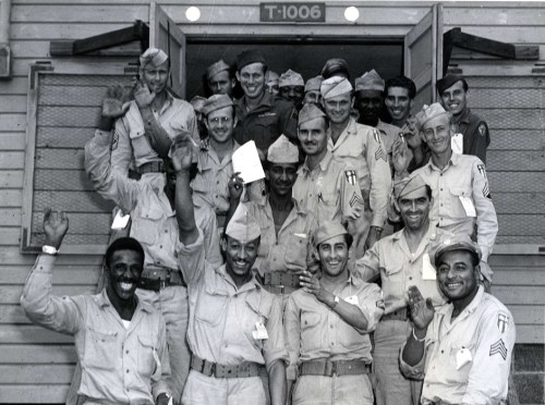 A group of soldiers in uniform smiling and waving outside a building, showcasing camaraderie and teamwork.