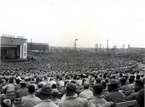 A large crowd gathers at a stadium, with a stage in the foreground and buildings in the background.