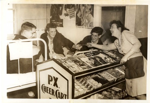 Four people interact around a magazine stand in a vintage setting, with posters on the wall and a bed in the background.