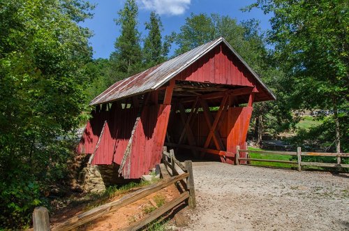 A red covered bridge with a slanted roof, surrounded by trees and a gravel path, under a blue sky.