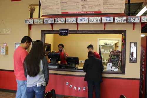 A busy restaurant counter with customers ordering from staff, featuring a menu and decorative license plates on the wall.