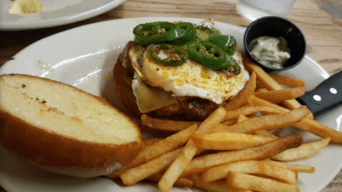 A sandwich with a fried egg, jalapeños, and cheese, served with a side of fries and a small cup of sauce.