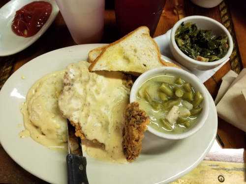 A plate of fried chicken smothered in gravy, served with toast, collard greens, and a bowl of green beans.