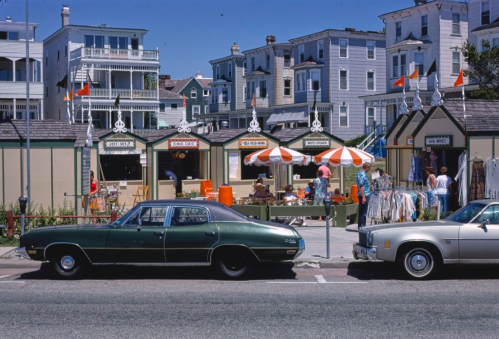 A vintage scene featuring two classic cars parked in front of colorful beach shops and people browsing outside.