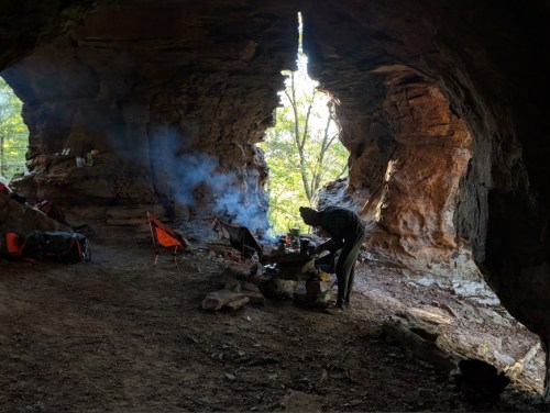 A person cooking at a campsite inside a rocky cave, with smoke rising and sunlight filtering through the opening.
