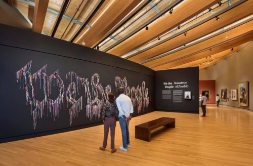 Visitors admire an art installation featuring colorful text on a black wall in a modern gallery space.