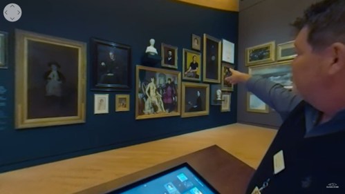 A person points at various framed artworks on a gallery wall, with a digital display in the foreground.