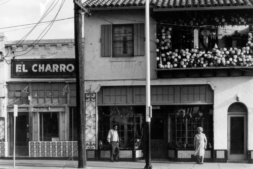 Black and white photo of a street scene featuring "El Charro" sign, decorated building, and two pedestrians.