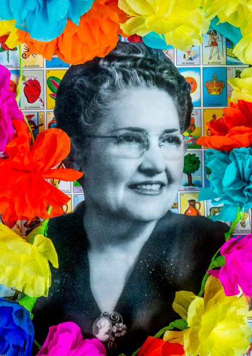 A black-and-white portrait of a woman with glasses, surrounded by colorful paper flowers and traditional game cards.