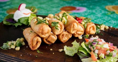 A plate of crispy rolled tacos garnished with herbs, served with fresh salad and salsa on a wooden board.