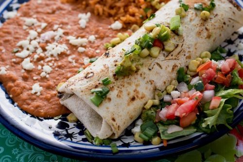 A plate featuring a burrito with toppings, accompanied by refried beans, rice, and a fresh salad with tomatoes and corn.