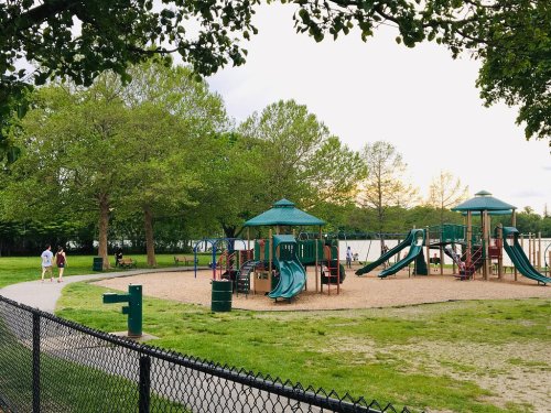 A playground with slides and swings surrounded by trees, with a walking path and a lake in the background.