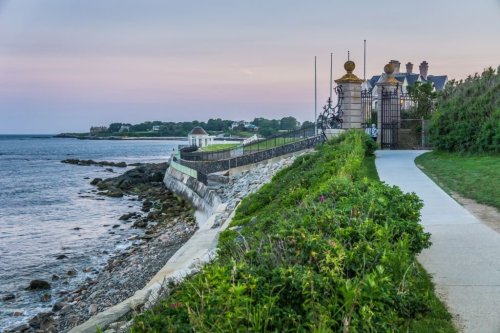 Coastal pathway with lush greenery, leading to ornate gates by the water, under a pastel sky at dusk.