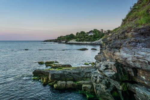 Coastal view at dusk, featuring rocky shoreline, calm water, and distant greenery under a pastel sky.