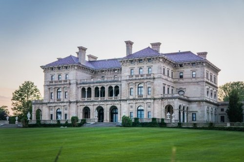A grand, historic mansion with intricate architecture, surrounded by lush green lawns and trees under a clear sky.