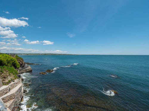 A serene coastal view with clear blue water, rocky shoreline, and a bright sky dotted with clouds.