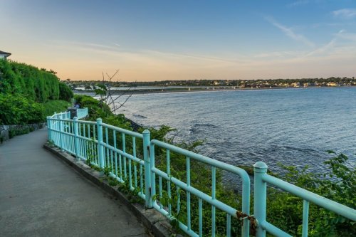 A scenic waterfront path with a turquoise railing, surrounded by greenery and a calm body of water under a clear sky.