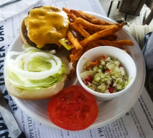 A plate with a cheeseburger, sweet potato fries, a slice of tomato, lettuce, and a small cup of coleslaw.