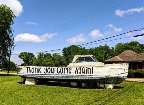 A boat on display in a grassy area with the words "Thank you! Come again!" painted on its side.