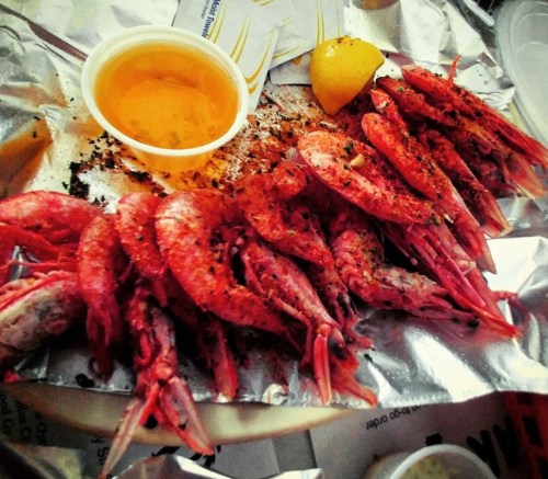 A plate of seasoned shrimp on foil, accompanied by a lemon wedge and a small cup of sauce.