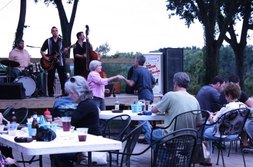A lively outdoor scene with a band playing, couples dancing, and people enjoying drinks at tables.