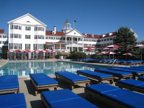 A large white hotel with a pool and blue lounge chairs, surrounded by red umbrellas and clear blue skies.