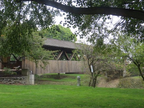 A wooden covered bridge surrounded by trees and green grass, with a stream visible in the background.