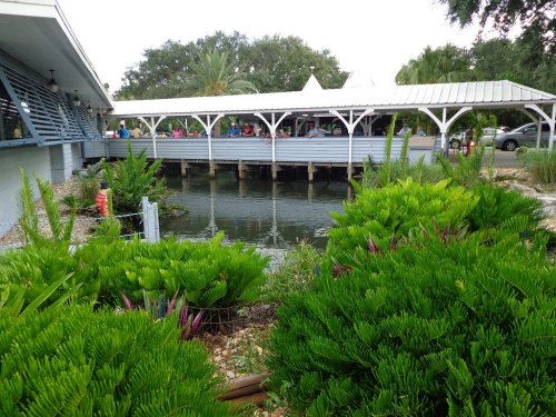 A waterfront restaurant with a wooden deck surrounded by lush greenery and a calm pond. People are dining outside.