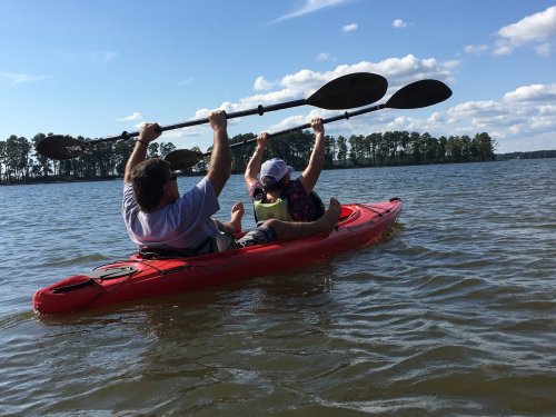 Two people in a red kayak paddle together on a calm lake, surrounded by trees and a blue sky.