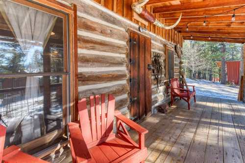 A rustic cabin porch with two red chairs, wooden logs, and a view of trees in the background.