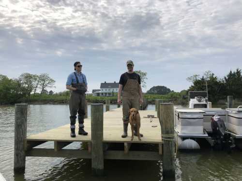 Two men in waders stand on a dock with a dog, surrounded by water and boats under a cloudy sky.