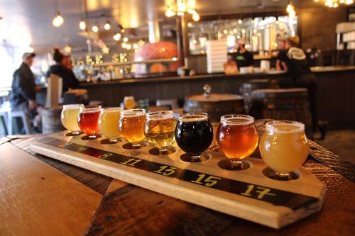 A wooden flight board with eight small glasses of various craft beers, set in a cozy brewery with warm lighting.