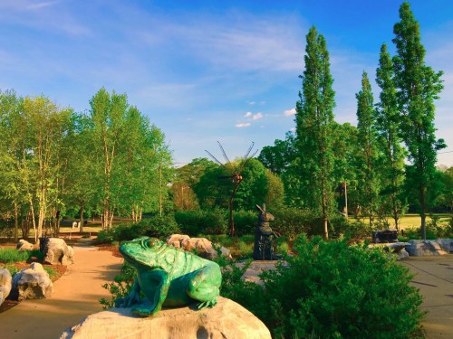 A serene park scene featuring a bronze frog sculpture, lush greenery, and tall trees under a clear blue sky.