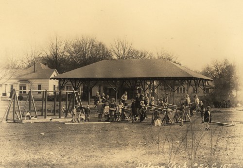 Children playing on wooden structures in Delano Park, with a pavilion and houses in the background. Vintage black and white photo.