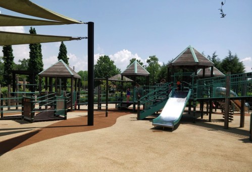 A playground with multiple structures, slides, and shaded areas, surrounded by trees and a clear blue sky.