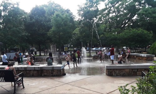 Children and adults play in a splash pad surrounded by trees on a sunny day. Water sprays from various fountains.