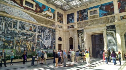 Visitors admire large murals in a spacious, well-lit gallery with intricate artwork on the walls.