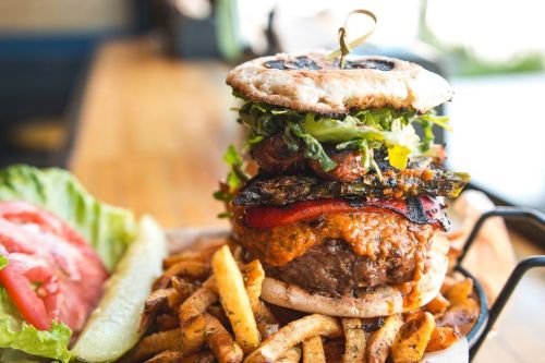 A towering burger with layers of lettuce, grilled vegetables, and sauce, served with fries and fresh vegetables on the side.