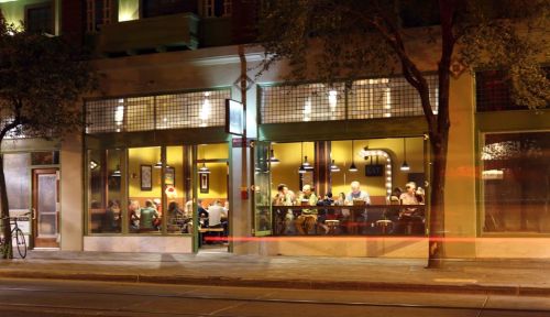 A bustling restaurant at night, with diners visible through large windows and warm yellow lighting.